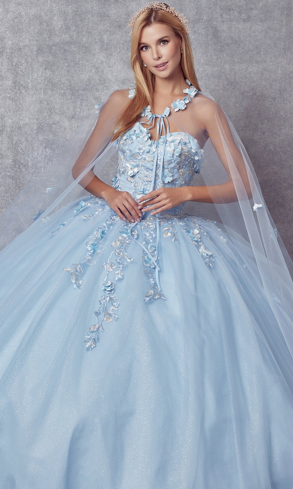A woman in the Juliet 1435 Strapless Ball Gown Quinceañera Dress, featuring light blue embroidery, floral embellishments, and a sheer cape, stands before a gray textured background.